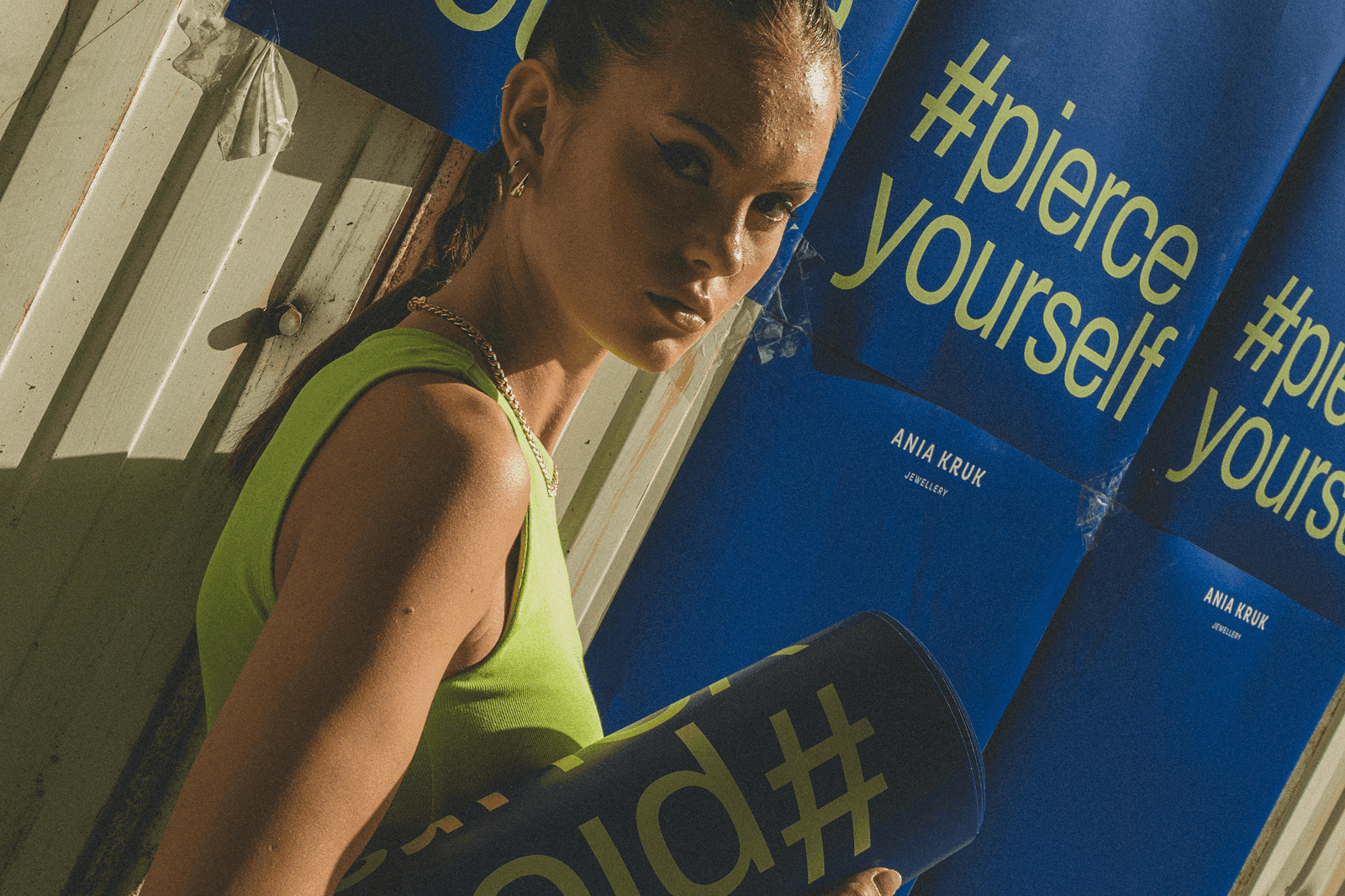 Kobieta ubrana w jasnozielony top, prezentująca kolczyki Ania Kruk na tle ściany z napisem zawierającym hashtag #pierceyourself.